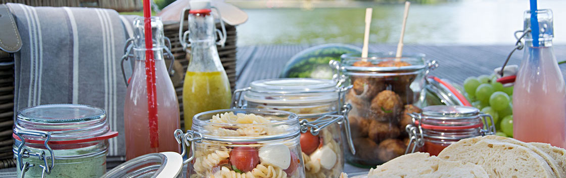 Picknickkorb, davor Gläser mit Nudelsalat und Fleischbällchen und Flaschen mit Erfrischungsgetränken